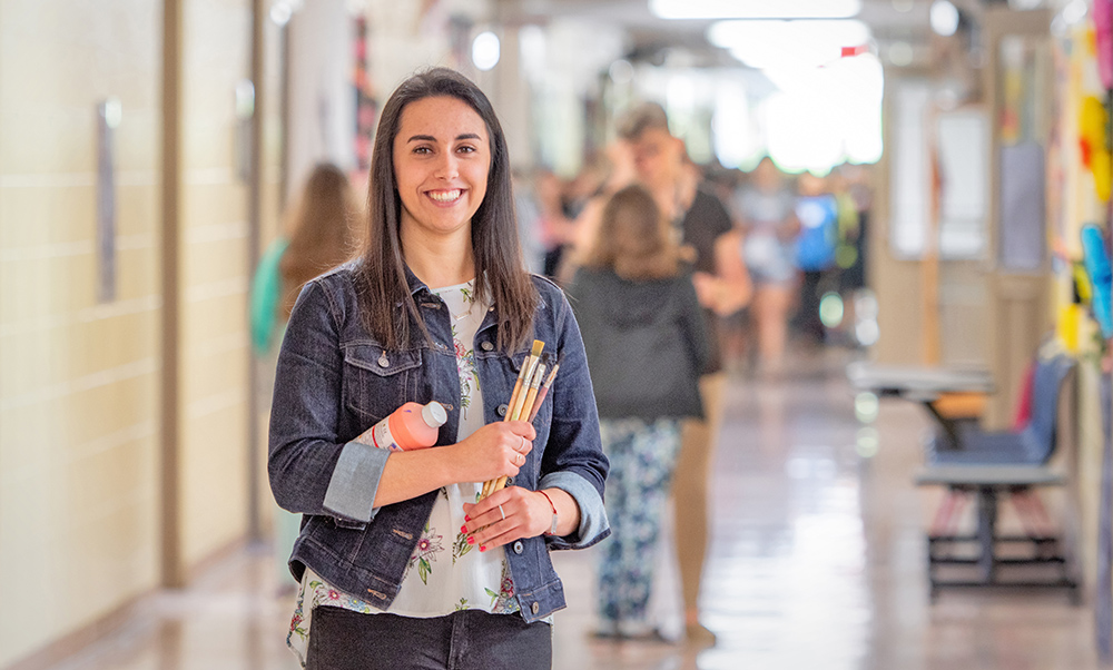 female MSJ student teacher in hallway holding paint and brushes.